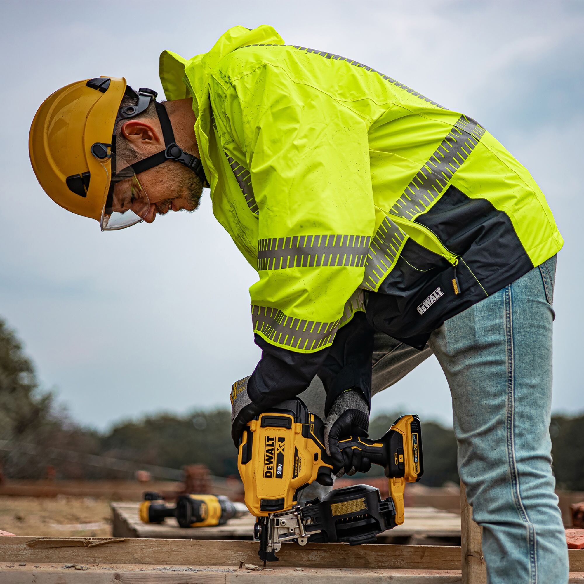 WORKER IN A DRW21 HI-VIS GREEN RAINJACKET AND YELLOW HELMET USING A DEWALT POWER TOOL ON A WORKSITE, FOCUSING ON WOOD FRAMING